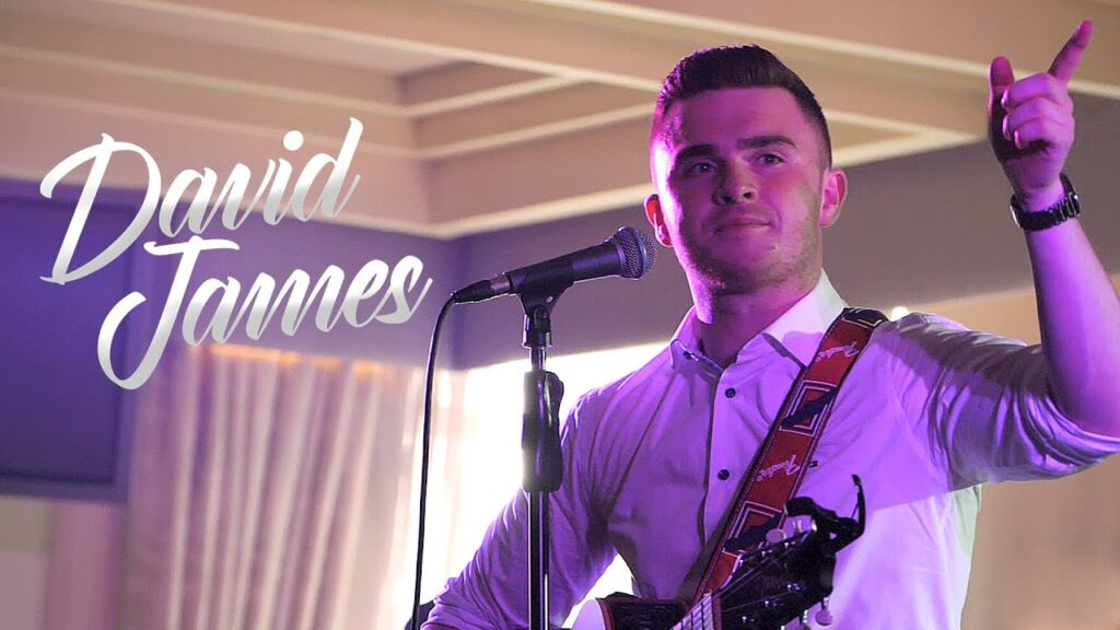 Promotional image of singer David James performing live on stage with an acoustic guitar and microphone.